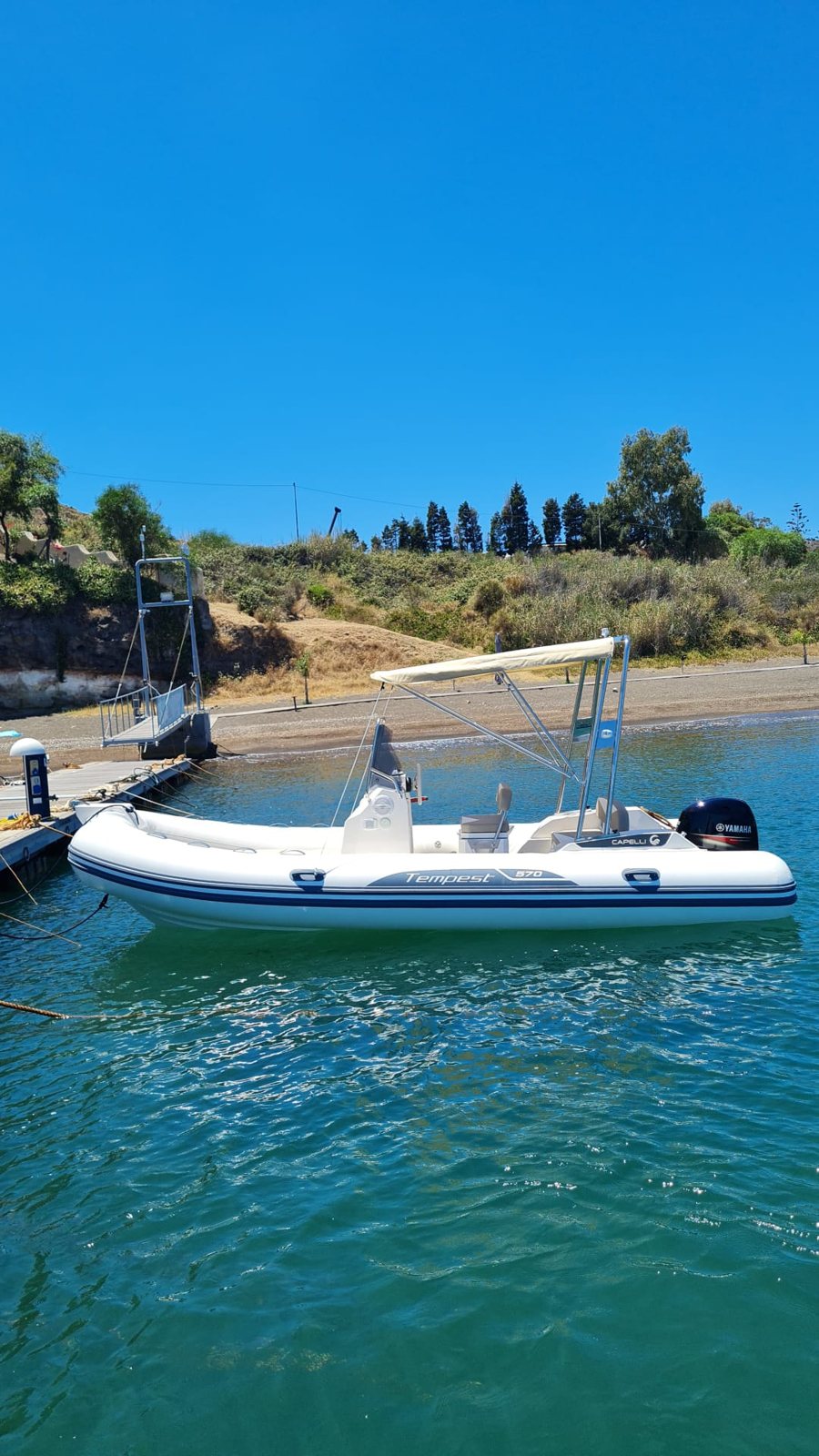 Gommone bianco Capelli Tempest 570 con tendalino parasole e motore Yamaha, ormeggiato al pontile galleggiante; dietro, la spiaggia scura e la collina